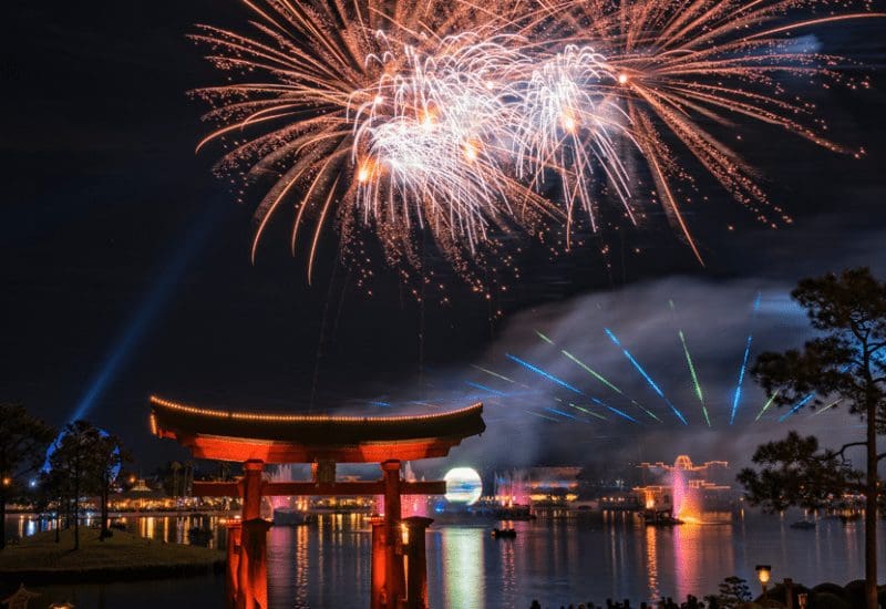 Fireworks illuminate the night sky above a tori gate, casting a vibrant reflection on the water.