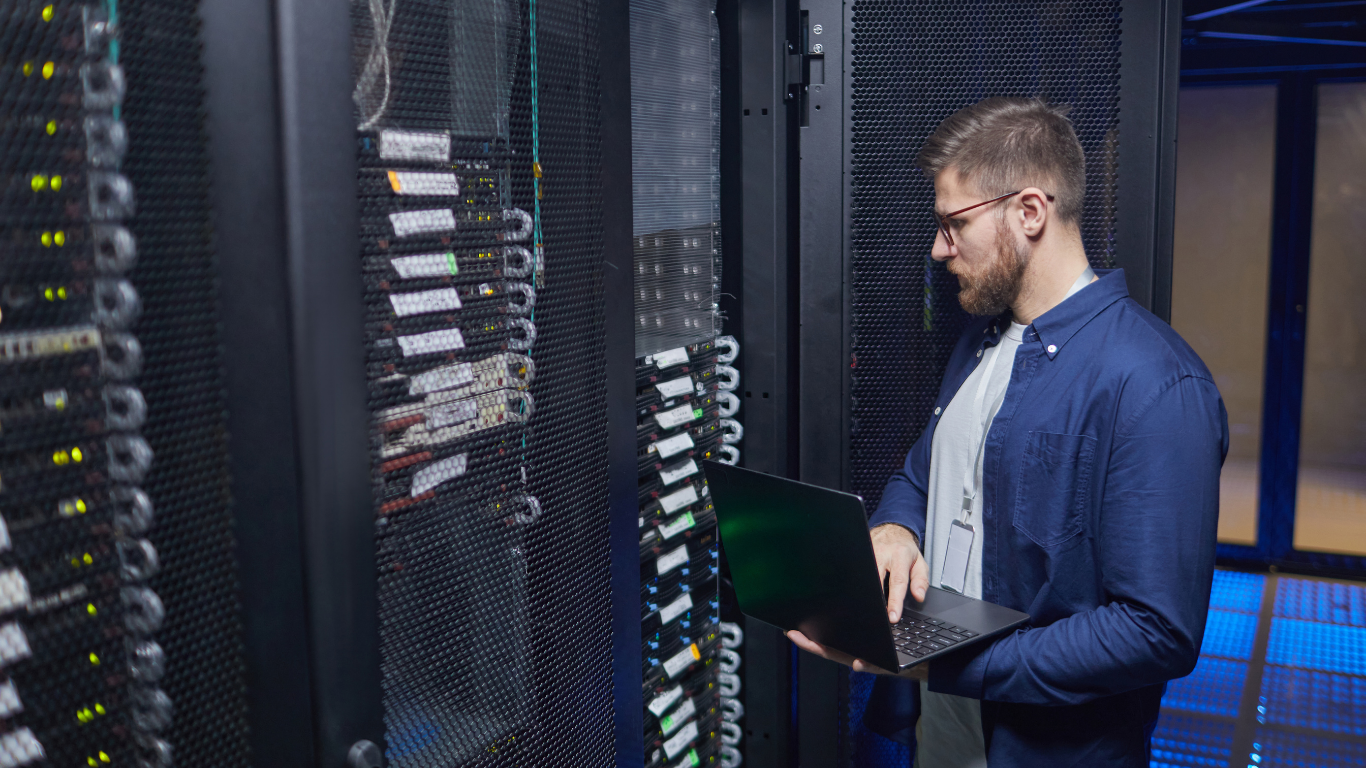 An IT technician with a beard and glasses using a laptop while inspecting server racks in a data center.